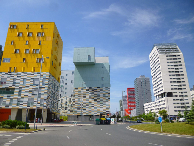 Eine Stadtstraße mit hohen, einzigartig gestalteten Universitätsgebäuden im Hintergrund, Straßenlaternen, Straßenbeleuchtung, fahrende Fahrzeuge, Informationsschilder, Grünflächen und eine bewölkte Himmel.