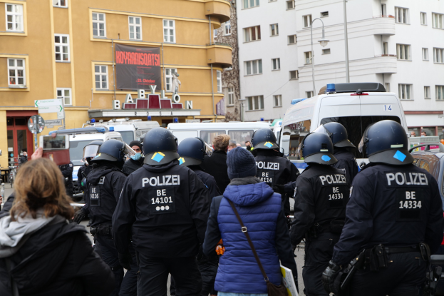 Polizeibeamte in Uniform vor einer Menge während einer Demonstration in Berlin, mit Fahrzeugen, Gebäuden und einer Kamera im Bild.