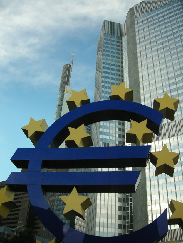 Das Euro-Zeichen steht prominent vor dem Hauptquartier der Europäischen Zentralbank, einem großen modernen Gebäude, das von Bäumen umgeben ist.