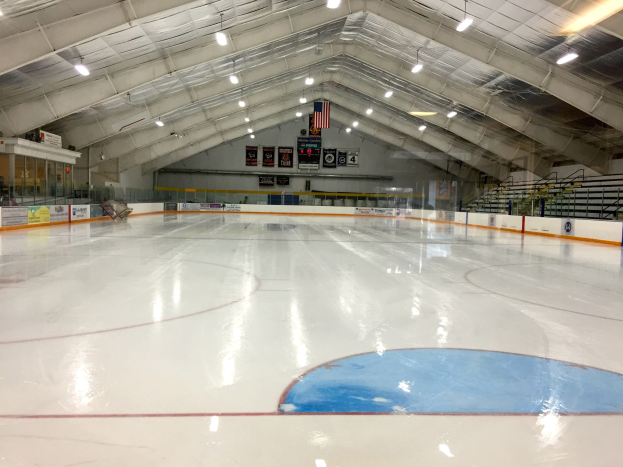 Große Indoor-Eisfläche mit Deckenbeleuchtung, Werbetafeln, einer Rückwand, Treppen mit Geländern und einigen Menschen, die einem Hockey-Sportplatz ähneln.