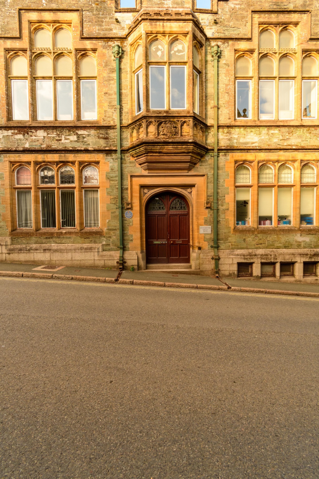 Altes Gebäude mit vielen Fenstern und einer hölzernen Tür, das neben einer Straße steht und an dessen Wänden Rohre verlaufen.