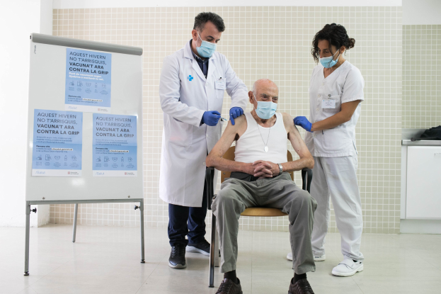 Alter Mann sitzt in einem Stuhl und bekommt von zwei maskierten Ärzten in einem Krankenhauszimmer eine Impfung verabreicht, wobei ein Schild und Gegenstände in der Nähe zu sehen sind.