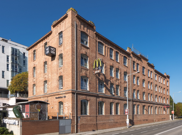 Ein großes Backsteingebäude von McDonald's an einer Straßenecke in Berlin, Deutschland, mit Straßenlaternen, Schildern, Fahrzeugen, Grünflächen und einem bewölkten Himmel.