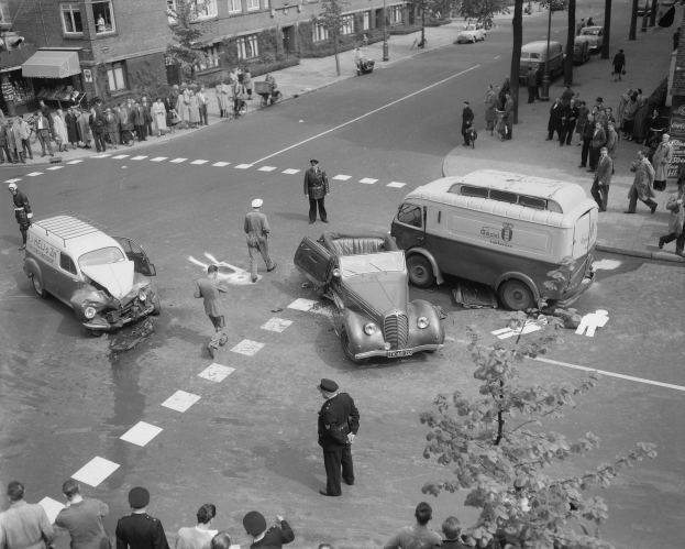 Schwarz-weißes Bild eines Autounfalls an einer Straßenkreuzung mit mehreren Fahrzeugen, darunter ein Lieferwagen, und einer Gruppe von Zuschauern; Gebäude, Bäume, Strommasten und Texttafeln sind im Hintergrund sichtbar.