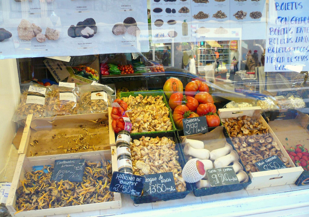Marktstand-Vitrine mit Gemüse, Flaschen und anderen Lebensmitteln, Papieren mit Text auf der Scheibe und Menschen im Hintergrund.