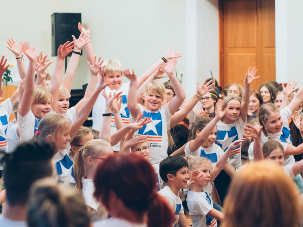 Kinder mit erhobenen Händen vor einer Menge, in der Nähe einer Wand und einer Tür, auf einem Sommercamp in Schweden mit einem Tisch, einer Vase und einem Lautsprecher links daneben.