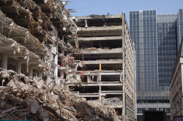 Ein großes Gebäude wird in der Innenstadt abgerissen, umgeben von intakten Strukturen, Straßeninfrastruktur, Fahrzeugen, Bäumen und einem klaren Himmel.