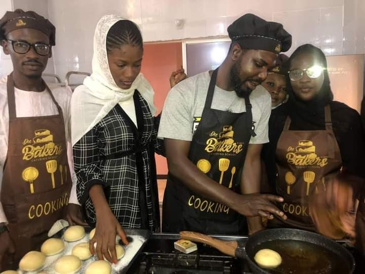 Eine Gruppe von Menschen in Schürzen und Mützen bereitet in einer Küche Essen zu, umgeben von einem Herd mit einem Topf Essen, mit Schränken und einer Wand im Hintergrund.