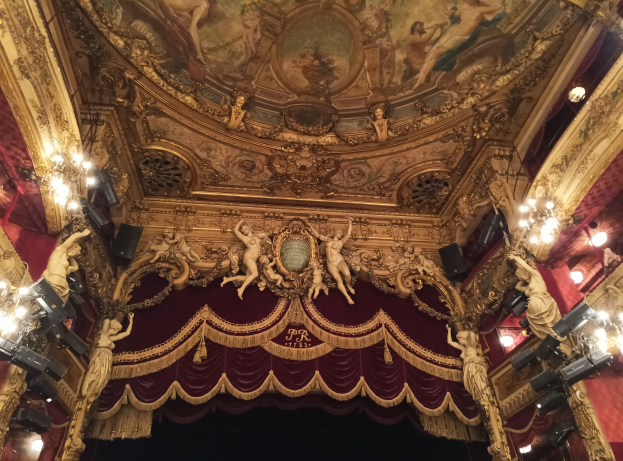 Innensicht eines Theaters mit roter Gardine, Kronleuchtern, Skulpturen, Lichtern und einer bemalten Decke, die eine Bühne und einige Personen am Boden zeigt.