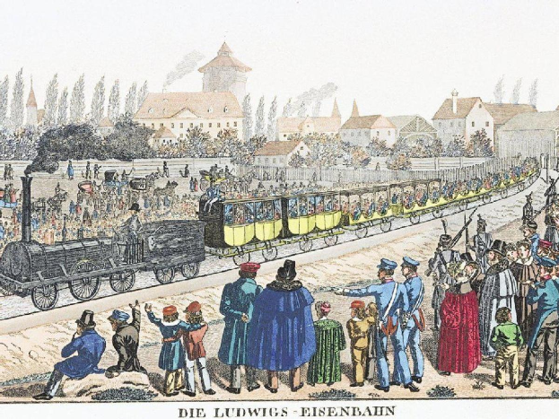 Eine Gruppe von Menschen in der Nähe eines Zuges auf Schienen stehend, einige sitzen im Zug und andere auf dem Boden, vor Gebäuden und Bäumen mit Text "Ludwigs-Eisenbahn, die erste Bahnstation in Deutschland" unten.