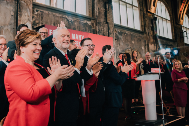 Eine Gruppe von Menschen, die vor einem Publikum applaudieren, mit einem Podium, einem Mikrofon und einer Texttafel rechts und Stühlen, einem Banner, einer Wand, Fenstern und Lichtern im Hintergrund.
