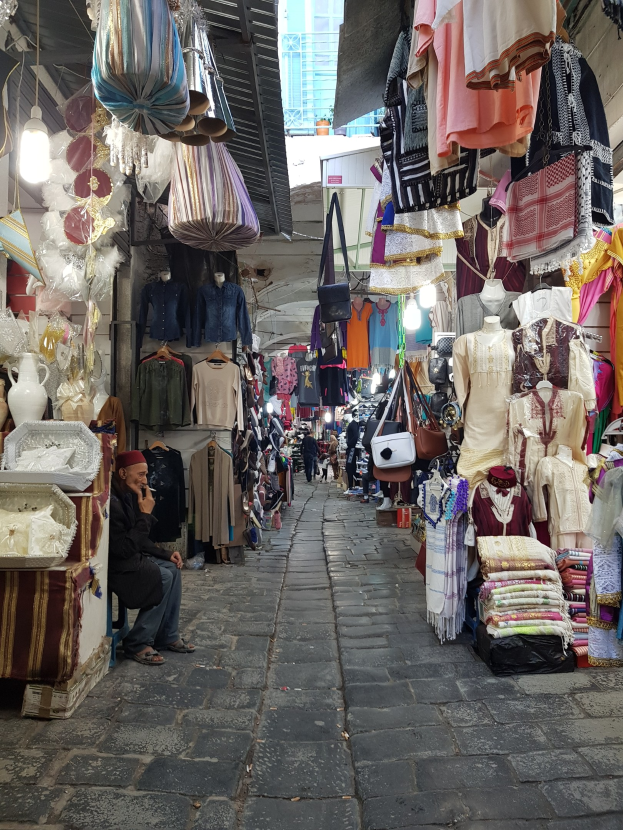 Ein belebter Straßenmarkt in Jerusalems Altstadt mit Ständen, die Kleidung, Taschen, Lampen und andere Artikel verkaufen, während Menschen umhergehen und die Stände durchstöbern, mit einem Gebäude im Hintergrund.