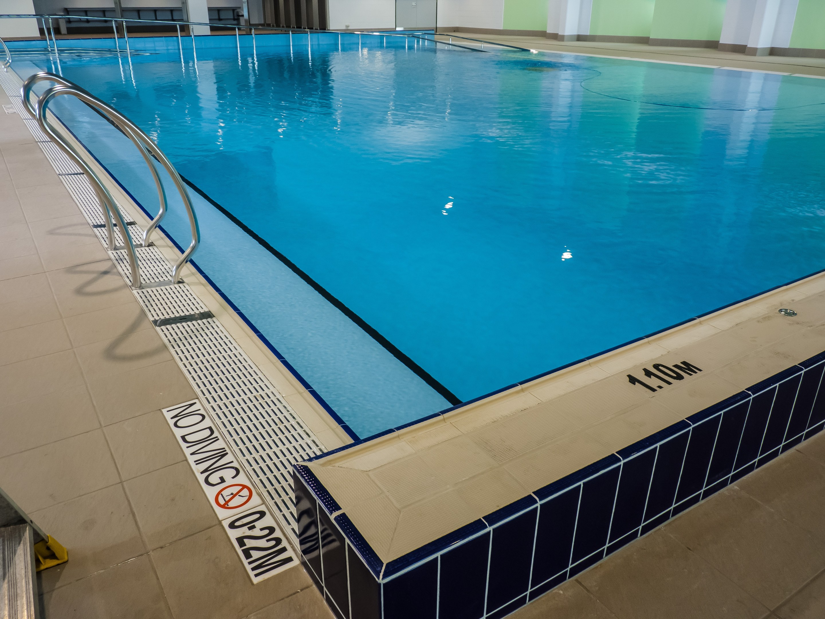 Großes Hallenbad in einem geräumigen Gebäude mit gefliestem Boden, Metallträgern, einer Wandtafel, einer Tür und Deckenbeleuchtung.