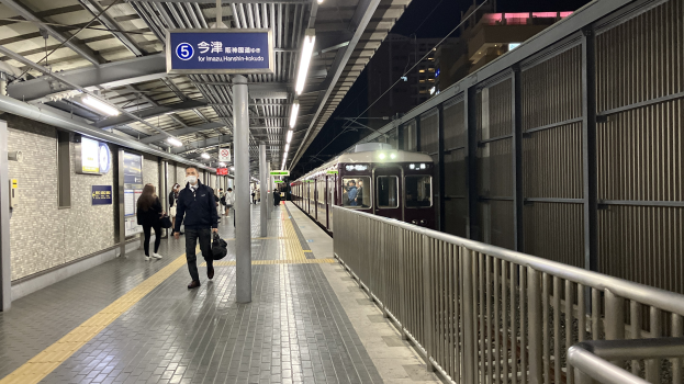 Ein Zug fährt auf einen Bahnsteig ein, mit Menschen in der Nähe, einem Geländer auf der rechten Seite, Gebäuden im Hintergrund, Bahnsteiglampen und einer Abfahrtsanzeige.