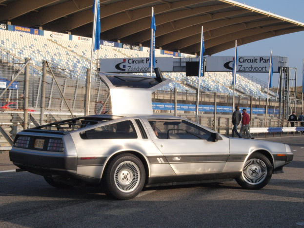 Ein silberner DeLorean DMC-12 fährt auf der Straße, umgeben von Menschen, Fahnen, Schildern, Geländern, Stühlen und einem Schuppen, mit Bäumen und einem klaren blauen Himmel im Hintergrund.
