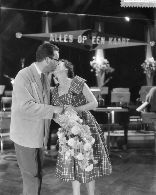 Ein Mann und eine Frau umarmen und küssen sich auf einer Bühne, während die Frau einen Blumenstrauß hält, mit Stühlen und Blumenvasen im Hintergrund.