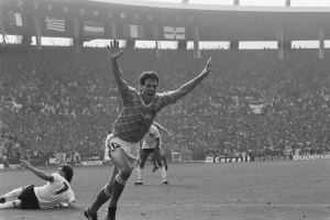 Schwarzes und weißes Foto eines Fußballspielers, der ein Tor feiert, umgeben von Teamkollegen und Stab auf dem Feld mit Stadiontafeln, Fahnen und Pfosten im Hintergrund.