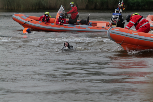 Eine Gruppe von Menschen in einem aufblasbaren Boot auf einem Fluss, mit zwei Personen im Wasser im Vordergrund und Vegetation im Hintergrund; alle tragen Schwimmwesten und Helme, was auf einen Rettungseinsatz hindeutet.