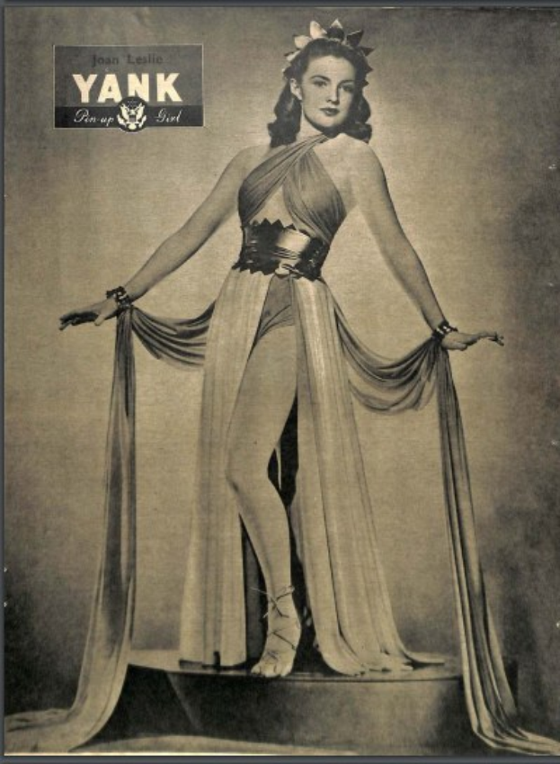 Ein Schwarz-Weiß-Foto von Schauspielerin Joan Leslie in einem langen, hochgeschlossenen, langärmligen Kleid mit einem klassischen 1940er-Stil-Haarschnitt und dezentem Make-up, die selbstbewusst auf einer Bühne steht.