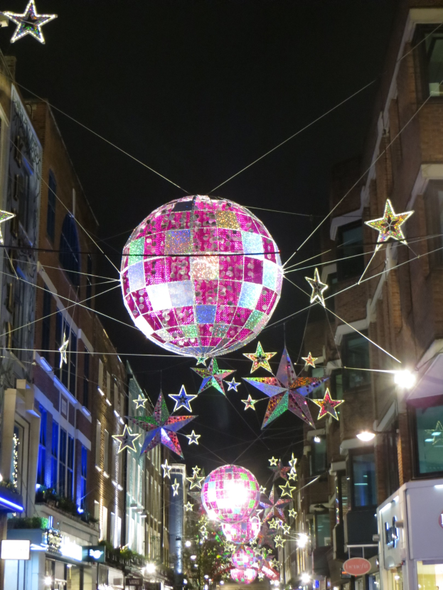 Eine festlich geschm├╝ckte Stra├če in einer Stadt bei Nacht mit zahlreichen Lichtern und Deko, mit einer gro├čen Diskokugel, die von einem Geb├Ąude h├Ąngt.