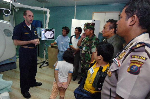 Eine Gruppe von Menschen in einem Krankenhauszimmer mit einem Mann, der ein Mikrofon hält, einem Bett mit einer Decke, medizinischen Geräten, einem Röntgengerät, einer Texttafel, einer Tür und Deckenleuchten.