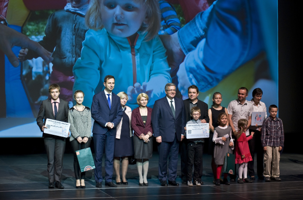 Eine Gruppe von Menschen auf einer Bühne mit Taschen und Urkunden, mit einem Bildschirm im Hintergrund, der ein Bild eines Kindes und andere zeigt.