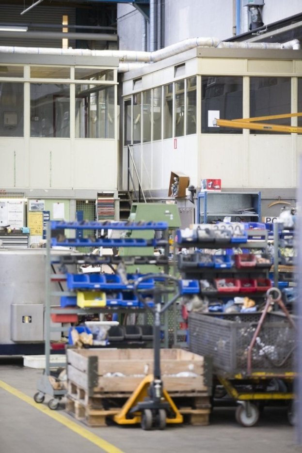 Ein großer Lagerraum voller verschiedener Ausrüstungen wie Wagen, Schränken und einer Kiste, mit einer Wand aus Glasfenstern und Rohren im Hintergrund.