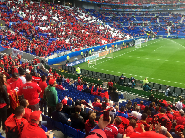 Eine große Menschenmenge in einem Stadion schaut einem Fußballspiel zu, mit Sitzplätzen und Stehplätzen, Fahnen, einem Torpfosten und einer Überdachung mit Deckenleuchten.