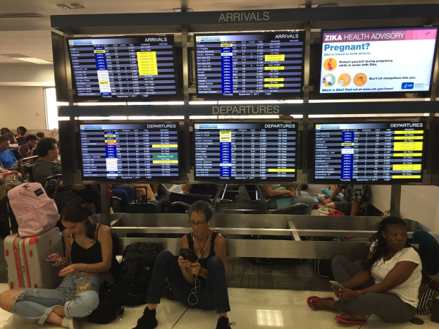 Gruppe von Menschen auf einem Flughafenboden sitzend, einige halten Handys, mit verstreuten Taschen und Bildschirmen mit Zika-Virus-Information im Hintergrund.