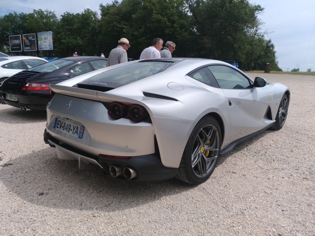Roter Ferrari 812 Superfast beim Goodwood Festival of Speed, umgeben von Menschen mit Mützen, Bäumen, Texttafeln und bewölktem Himmel im Hintergrund.