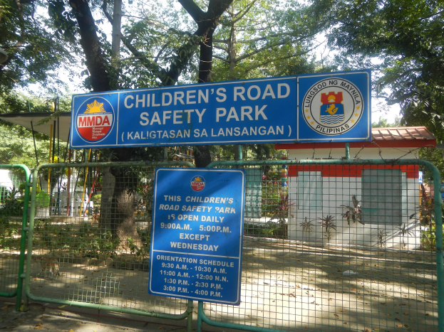 Kindersicherheitspark mit Metallzaun, zwei blauen Informationsschildern, einem kleinen Haus, Pflanzen, Bäumen und einem bewölktem Himmel.