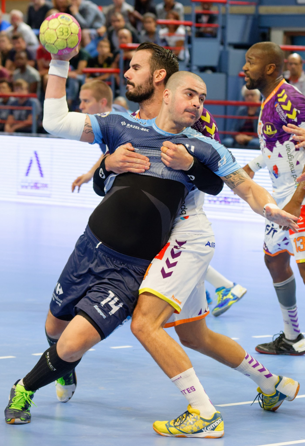 Eine Gruppe von Männern, die Handball in einem Stadion spielen, mit einem, der den Ball hält, und Zuschauern im Hintergrund.