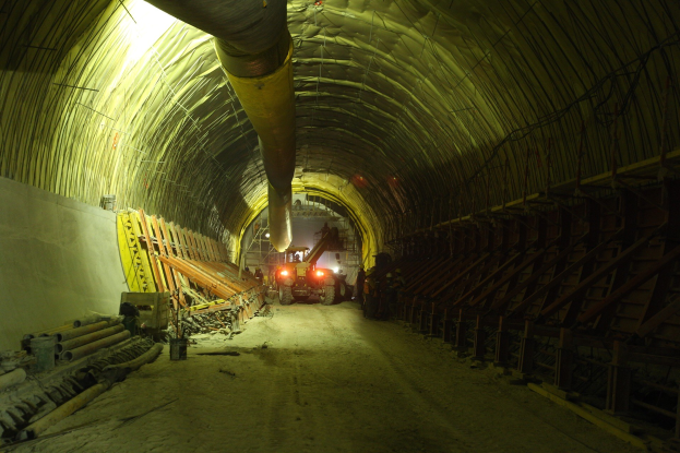 Großer Tunnel an einer Baustelle mit Fahrzeugen, verstreuten Holzobjekten, Rohren und einer Wand auf der linken Seite, beleuchtet von Deckenlampen.
