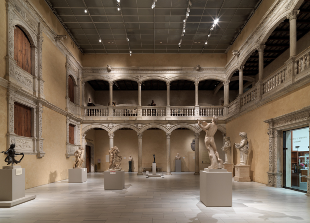 Ein geräumiger Museumsgalerie mit zahlreichen Statuen und Skulpturen auf Sockeln, mit architektonischen Elementen wie Wänden, Säulen, Geländern und Deckenleuchten und einigen Besuchern, die die Kunstwerke betrachten.