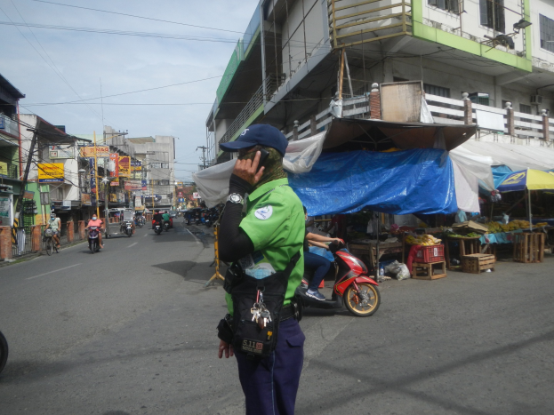 Ein Polizist in grüner und blauer Uniform mit blauer Mütze steht an einer Straßenseite, spricht in ein Handy, mit Fahrzeugen auf der Straße, Marktbuden rechts von ihm, einem Gebäude mit Fenstern im Hintergrund und einem bewölktem Himmel.