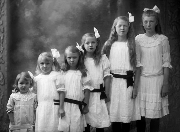 Eine Gruppe junger Mädchen in weißen Kleidern, die in einer Reihe vor einer Wand in einem Schwarz-Weiß-Foto stehen.