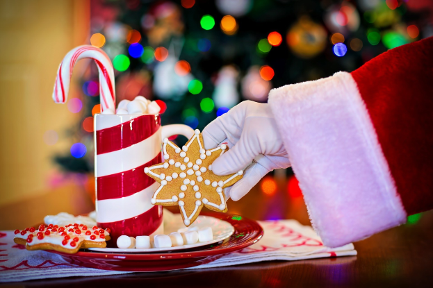 Eine Person mit einem Santa Claus Hut, die einen Becher heißer Schokolade und einen Bonbonstab hält und ein Teller mit Keksen und ein Tuch auf dem Tisch vor sich hat, vor einem Weihnachtsbaum und einer Wand.