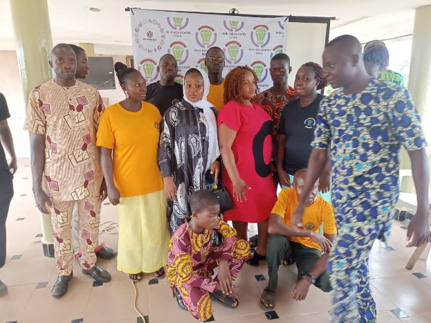 Gruppe von Menschen vor einem Banner mit der Aufschrift "Advocates in Ghana: Ein Schritt-für-Schritt-Führer" mit Stühlen, Säulen, einem Fernseher und einer Decke mit Deckenleuchten im Hintergrund.