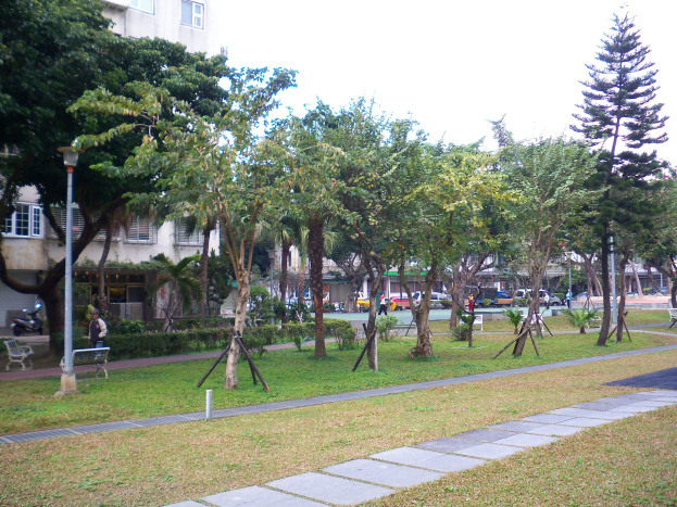 Ein Park mit grünem Gras, Bäumen, Bänken entlang eines Weges, Laternenmasten, Menschen, Fahrzeugen auf einer Straße, Gebäuden im Hintergrund und einem klaren blauen Himmel.