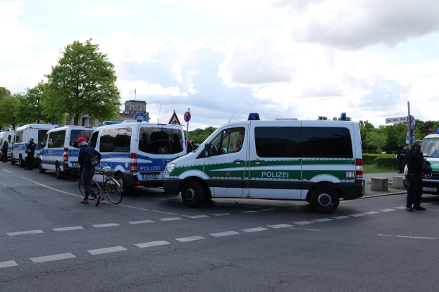 Eine Gruppe von Polizeiwagen, die entlang einer Straße geparkt sind, mit einem Radfahrer im Vordergrund und Gebäuden, Bäumen, Schildern im Hintergrund unter einem bewölkten Himmel.