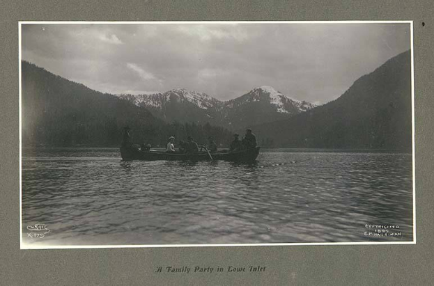 Schwarze und weiße Fotografie einer Familie in einem Kanu auf einem See mit Bergen und Bäumen im Hintergrund, Himmel sichtbar und Text unten.