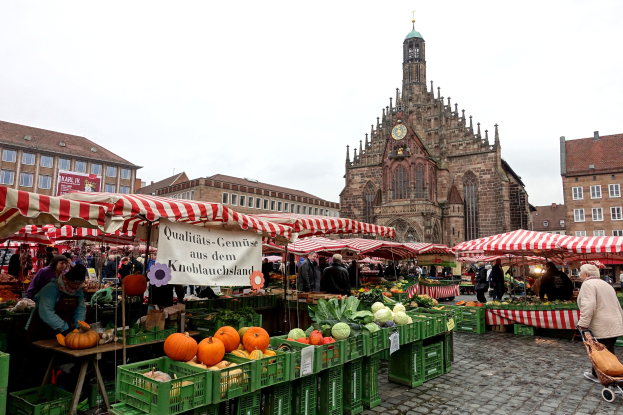 Ein belebter Markt in Nürnberg, Deutschland, mit farbenfrohen Obst- und Gemüsesorten auf den Ständen, Menschen mit Taschen und Zelten, vor einem Hintergrund aus Gebäuden mit Fenstern und einem Uhrenturm unter einem klaren Himmel.