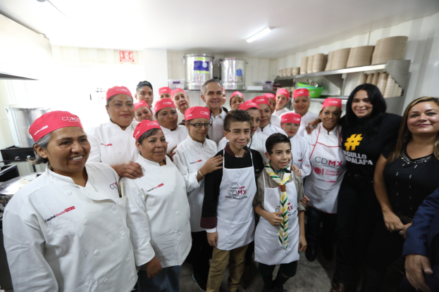 Eine Gruppe von Menschen, die in einer Küche lächeln und posieren, mit Utensilien links, Gestellen mit Tellern und Schüsseln im Hintergrund und Deckenlampen, was auf eine Feier einer Kochschule in Mexiko hindeutet.