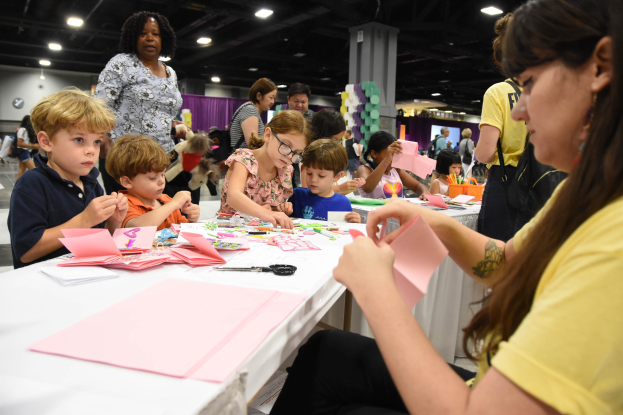 Eine Gruppe von Kindern und eine Frau in einem gelben Shirt sitzen an einem Tisch mit Bastelmaterialien während einer Bastelmesse, mit Menschen und Säulen im Hintergrund.