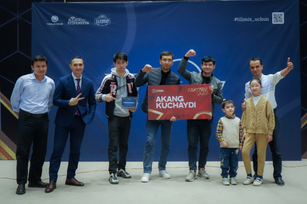 Gruppe von Menschen auf einer Bühne mit einer Tafel, auf der "Akang Kuchaydi - Zakovat Quiz" steht, mit einem Banner, Scheinwerfern und anderen Gegenständen im Hintergrund.