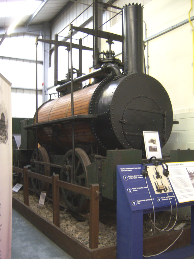 Alte Dampflokomotive in einem Museum mit Informationsschildern, einem Zaun und Deckenbeleuchtung ausgestellt.