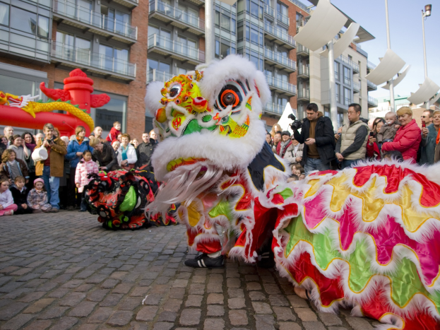 Ein lebendiges chinesisches Neujahrsfest in Amsterdam mit einem Löwen tanzen im Vordergrund und einer Menge Menschen drumherum, einige halten Kameras, vor Häusern, Laternenmasten und einem klaren blauen Himmel.
