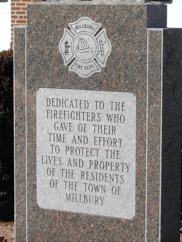 Denkmalstein mit einer Plakette, die Feuerwehren gewidmet ist, vor einem Backsteingeb├Ąude umgeben von Pflanzen und B├Ąumen.