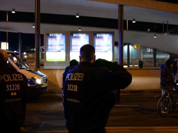 Eine Gruppe von Polizeibeamten steht vor einem Gebäude in der Nacht mit einem geparkten Auto auf der linken Seite und einem Radfahrer auf der rechten Seite, beleuchtet von Bildschirmen und Lichtern im Hintergrund.