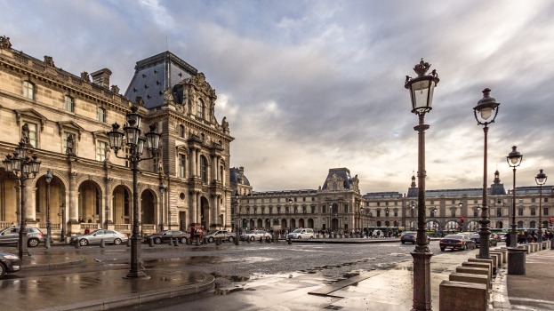 Außenansicht des Louvre-Museums in Paris, Frankreich, mit seiner ikonischen Architektur, Straßenlaternen, Lichtern, fahrenden Fahrzeugen, Fußgängern auf dem Gehweg und einer bewölkten Himmel.